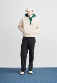 Beežist tõmblukuga bomberjakk rohelise kapuutsiga, kombineeritud tumedate teksade ja valgete vabaajajalanõudega, taustaks hele ühtlane sein.