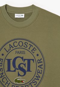 Felpa color verde oliva con un logo rotondo che riporta "LACOSTE," "PARIGI," e "ABBIGLIAMENTO SPORTIVO FRANCESE" in blu navy. Ricamo di coccodrillo verde.