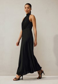 Schwarzes ärmelloses Kleid mit hohem Ausschnitt, drapiertem Oberteil und fließendem Rock. Kombiniert mit schwarzen, riemchenbesetzten High-Heel-Sandalen. Glatte Textur.