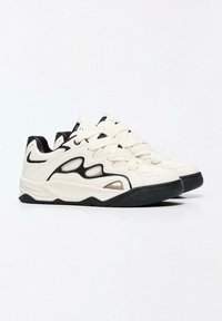 Fehér sportcipők fekete kiemelésekkel, texturált felsőrésszel, vastag fekete talppal és jellegzetes, kivágott dizájnelemekkel az oldalakon.