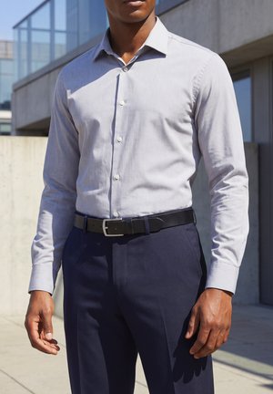 Man wearing light gray button-up shirt tucked into navy blue pants with black belt, standing outdoors near concrete building.