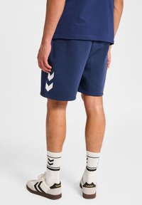 Man i marinblå shorts och matchande t-shirt, vita strumpor med svarta ränder och vita sneakers med svarta accenter.