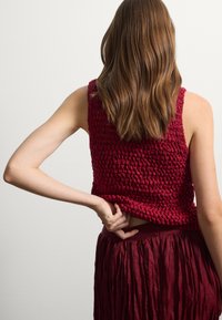 Top senza maniche bordeaux con design a maglia texturizzata. Il modello ha i capelli lunghi e indossa una gonna plissettata abbinata.