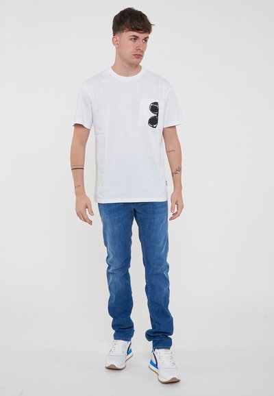T-shirt bianca in cotone con una stampa di occhiali da sole sulla tasca del petto, abbinata a jeans in denim blu e sneakers bianche.