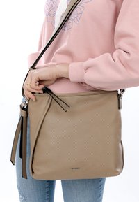 Isik, kes kannab roosat dressipluusi ja teksaseid, hoides käes beežikat perforatsioonidega nahast õlakotti, millel on lukk ja rihma detailid.