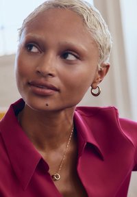 Majica s kratkimi rokavi magenta barve s ovratnikom, zlate obročeve uhane, nežno verižico z detajlom prepletenega obročka. Gladka tekstura kože.