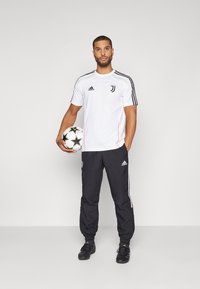 Biała sportowa koszulka z czarnymi akcentami i logo Juventusu, w połączeniu z czarnymi spodniami z białymi paskami, trzymając czarno-białą piłkę nożną.