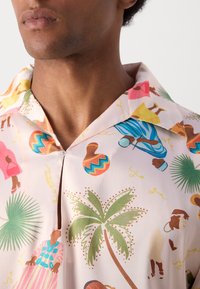 Camisa de manga corta de tela rosa claro con un estampado vibrante de figuras, palmeras y patrones en verde, marrón, amarillo y azul.