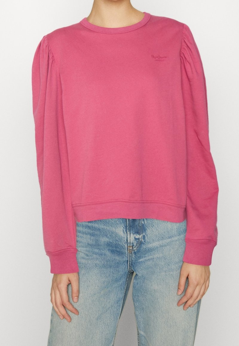 Sweatshirt rose avec manches bouffantes, col rond et ourlet côtelé. Confectionné dans un tissu doux, avec un petit logo sur la poitrine. Porté avec un jean bleu.