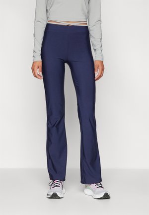Femme portant des leggings bleu marine ajustés évasés, un haut à manches longues gris clair et des chaussures de sport blanches avec des accents roses.