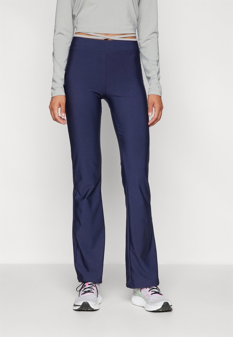 Femme portant des leggings bleu marine ajustés évasés, un haut à manches longues gris clair et des chaussures de sport blanches avec des accents roses.