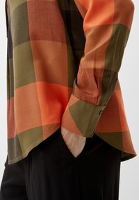 Camicia a maniche lunghe a quadri in tonalità arancioni, verdi e nere. Presenta un polsino con bottoni e una vestibilità comoda, con orlo dritto e texture morbida.