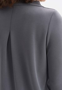 Top gris de manga larga con abertura en la parte trasera, textura suave y diseño minimalista. La tela tiene un acabado suave y ligeramente mate.