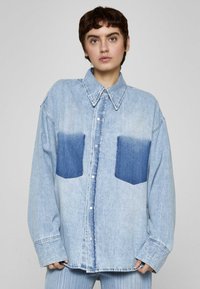 Camicia di denim azzurro chiaro con vestibilità comoda, caratterizzata da due tasche a toppa di un blu più scuro e colletto classico. Bottoni sulla parte anteriore.
