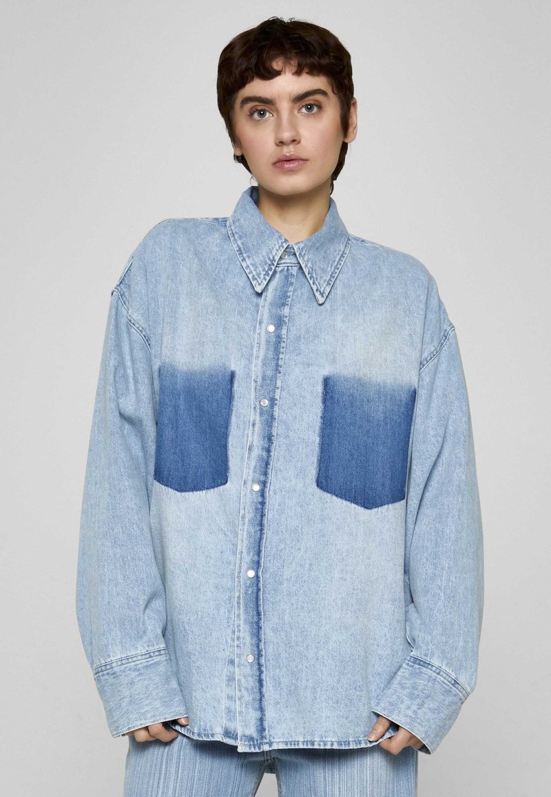 Camicia di denim azzurro chiaro con vestibilità comoda, caratterizzata da due tasche a toppa di un blu più scuro e colletto classico. Bottoni sulla parte anteriore.