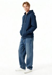 Felpa con zip di colore navy, dotata di tasche frontali, abbinata a jeans blu a vestibilità comoda e sneakers bianche. Linee pulite e design informale.