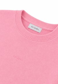 Gros plan d'un sweat-shirt rose à col rond côtelé avec le nom de la marque "OH APRIL" sur une étiquette blanche et embossé sur la poitrine.