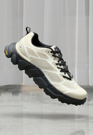 Chaussure de trail running beige clair et noire avec une semelle robuste, un dessus en mesh respirant et une languette à l'arrière, présentée flottant au-dessus d'un sol en marbre.