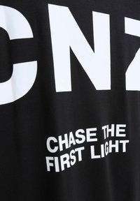 Camiseta negra con gran texto blanco "CNZ" y la frase "PERSEGUIR LA PRIMERA LUZ" en una fuente limpia y moderna. Suave textura de algodón.
