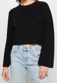 Schwarzer, gerippter, kurz geschnittener Pullover mit langen, ausgestellten Ärmeln. Kombiniert mit hellblauen, hoch taillierten Jeans mit lockerem Schnitt.