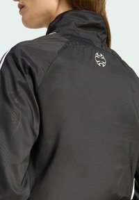 Giacca a vento nera con colletto alto, dotata di dettagli grafici sottili e un piccolo logo sul retro vicino alla scollatura.