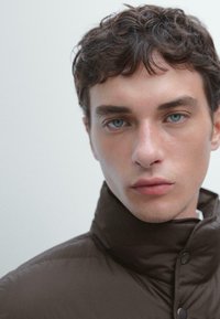 Giacca puffer marrone a collo alto con una texture liscia. Presenta chiusure a scatto e un design minimalista. La pelle del modello è chiara con occhi blu chiari.