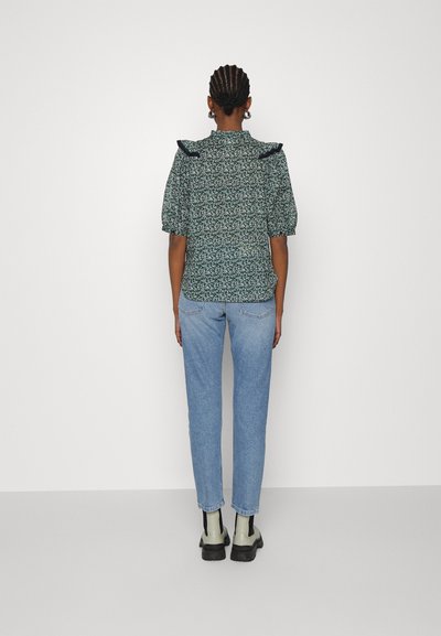 Μπλούζα με λουλουδάτο σχέδιο σε σκούρο πράσινο ύφασμα με φουσκωτά μανίκια και ριχτό γιακά, φορεμένη με ανοιχτό μπλε τζιν ίσιου διαστήματος και μαύρες μπότες.