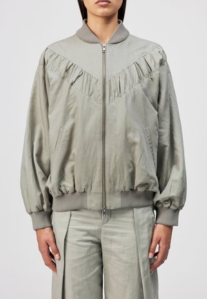 Frau trägt eine hellgraue Bomberjacke mit Reißverschluss, Rüschenbesatz am Einsatz und passenden lockeren Hosen vor einfarbigem Hintergrund.