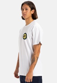 Camiseta de algodón blanca con cuello redondo y mangas cortas. Presenta un parche de logo redondo en amarillo, verde y negro en el pecho.