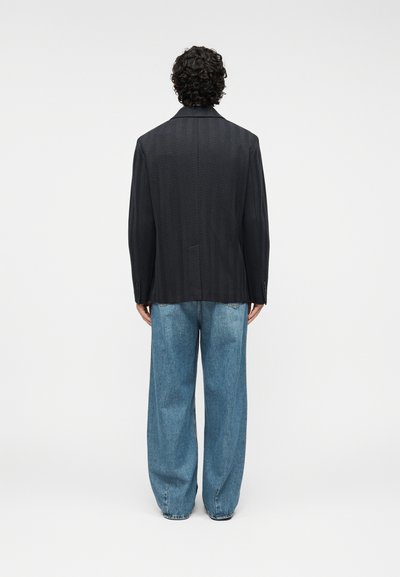 Veste noire texturée avec un motif en chevrons, associée à un jean en denim bleu à coupe ample. Coupe simple et moderne, sans éléments métalliques visibles.