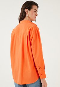 Oranje oversized shirt met lange mouwen, button-down kraag en een losse pasvorm. Gladde katoenen stof met een ontspannen achterontwerp en zijsplitten.
