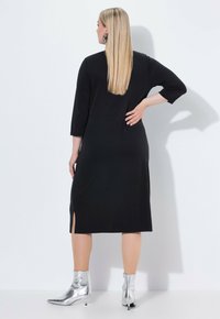 Vestido negro hasta la rodilla con mangas de tres cuartos, con aberturas laterales, combinado con botas brillantes plateadas hasta el tobillo. Textura de tela suave.