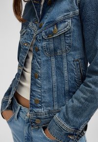 Close-up van een persoon die een blauwe Lee spijkerjas en spijkerbroek draagt, met de handen in de zakken, boven een witte cropped top.