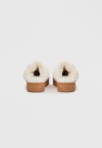 Pantofole fuzzy bianche senza lacci con suole in gomma beige, dotate di una foderatura spessa e morbida e dalla forma arrotondata. Nessun elemento metallico o motivo visibile.