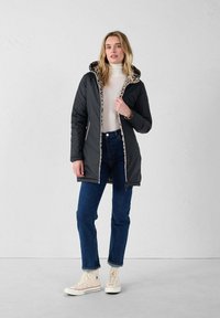 Chaqueta exterior acolchada negra con capucha, con un forro de estampado de leopardo y cierre de cremallera, combinada con vaqueros azules y zapatillas blancas.