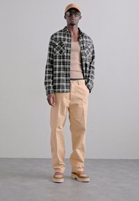 Rutet sort og hvid skjorte over en beige tank top, lysebeige bukser og beige sko med mønstrede stropper. Sorte solbriller og en ferskenfarvet kasket.