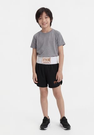 Enfant souriant debout portant un t-shirt gris, un short de boxe noir avec une ceinture blanche "Venum" et des baskets noires sur fond blanc.