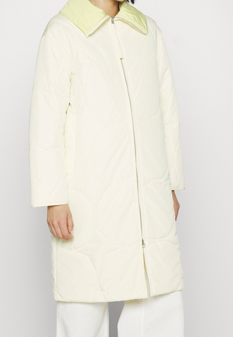 Abrigo acolchado crema de longitud media con cremallera frontal completa y collar verde pálido, llevado sobre pantalones blancos por una persona de pie.