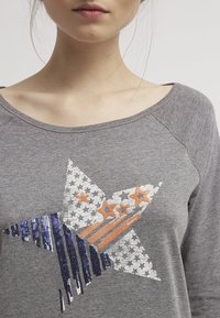 Grauer Sweatshirt mit einem großen Sternmuster, verziert mit Pailletten in Silber, Blau und Orange, und einem weiten, runden Ausschnitt.