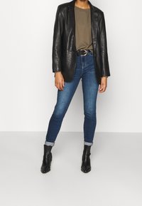 Blazer de cuero negro sobre una camiseta oliva, combinado con jeans ajustados azules y botines negros; texturas notables y diseño ajustado.