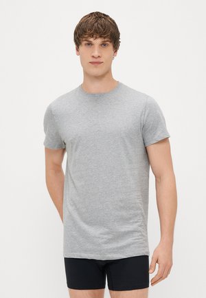 T-shirt cinza de manga curta, feito de algodão, com decote redondo e corte reto. Usado com calções pretos, em pé contra um fundo simples.