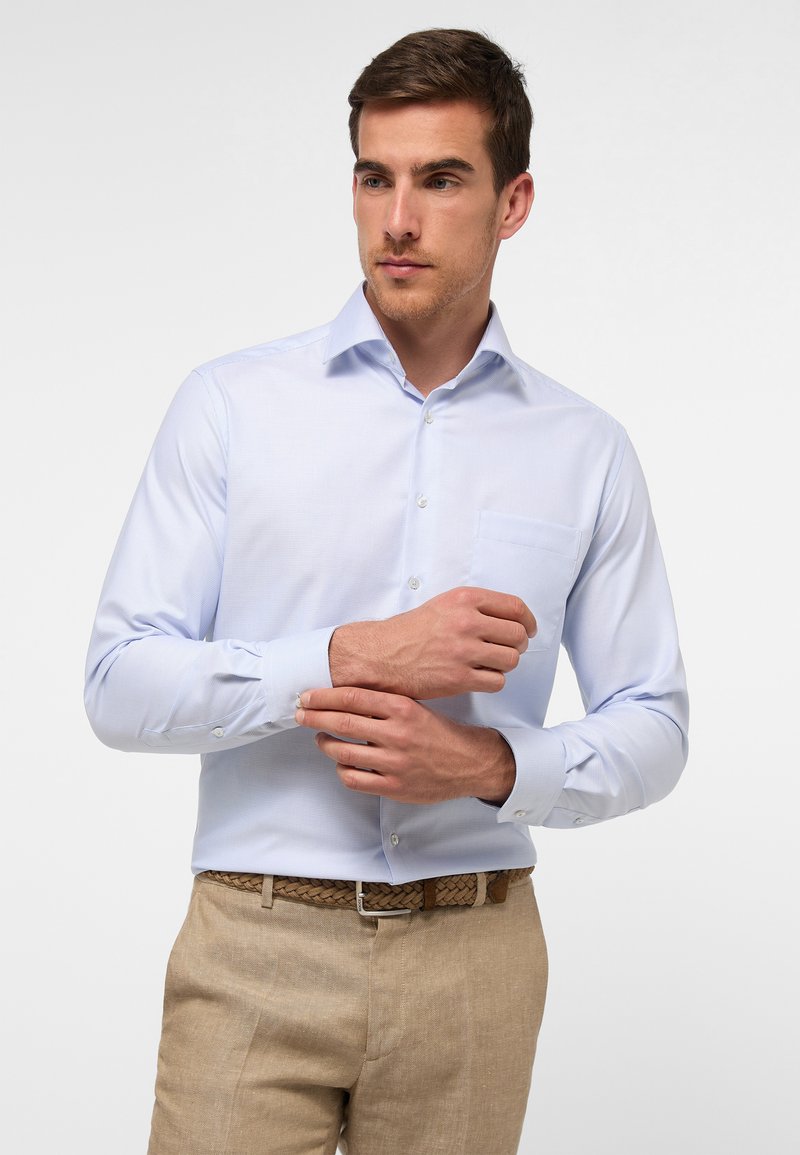Camicia azzurra chiara con colletto button-down, maniche lunghe, una tasca sul petto e una texture sottile, indossata con pantaloni beige.