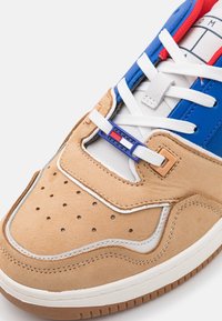 Zapatilla con parte superior de ante beige, acentos blancos, superposiciones de piel azul y un forro interior rojo. Presenta una caja de puntera perforada y un cierre de cordones con marca.