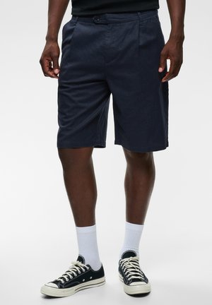 Mann trägt marineblaue Shorts, weiße Sportsocken und schwarze Sneakers mit niedrigem Schaft und steht vor einem einfarbigen Hintergrund.