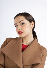 Barna, oversize kabát széles gallérral, sima textúrával, fényes piros blúzra rétegezve. A modellen egyenes, sötét haj és merész piros rúzs van.