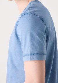 Blaues T-Shirt aus Baumwolle mit kurzen Ärmeln, Rundhalsausschnitt, sichtbaren Nahtdetails und glatter Textur, seitlich dargestellt.