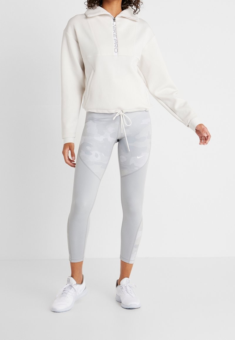 Kvinde iført en hvid hoodie med halvlukning, grå camouflage-mønstrede leggings med snøre i taljen og hvide træningssko på en ensfarvet baggrund.