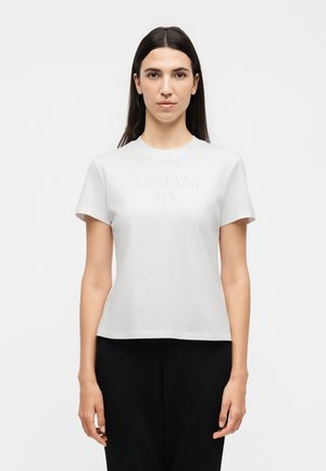 Femme aux longs cheveux foncés portant un t-shirt blanc Armani A|X à manches courtes et un pantalon noir, debout devant un fond blanc uni.