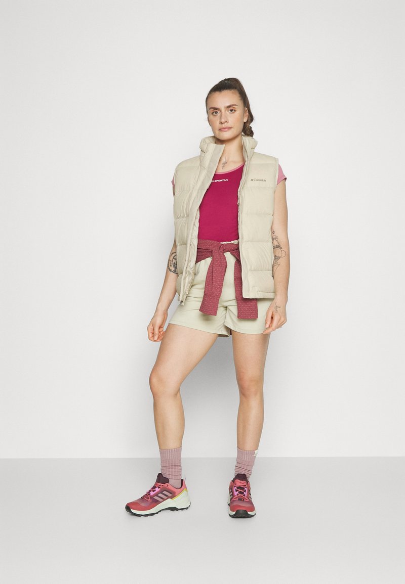 Leichter beiger Puffer-Weste über einem pinken Oberteil, kombiniert mit hellgrünen Shorts und pinken Sportschuhen. Auffällige Details sind ein gebundeter roter Akzent.