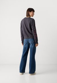 Felpa grigia scura con motivi colorati sulle maniche, abbinata a jeans blu larghi e scarpe chiare. Tessuto morbido, vestibilità rilassata.